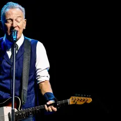 Bruce Springsteen z gitarą na scenie, w niebieskiej kamizelce i białej koszuli. Niezapomniane koncerty legendy rocka.