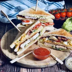 Leckere Sandwich-Ideen: Gestapelte Club-Sandwiches mit Hähnchen, Speck, Ei und Tomaten auf einem Holzbrett, daneben Tomaten und Salat.
