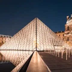 Piramide Luwru nocą z odbiciem w wodzie i oświetlonymi budynkami muzeum.