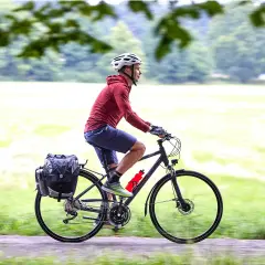 Mężczyzna w kasku jedzie na rowerze trekkingowym z sakwami, ciesząc się przejażdżką pośród zieleni.