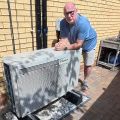 Un homme pose près d'une unité extérieure de pompe à chaleur Daikin, un système efficace pour le chauffage et la climatisation, potentiellement alimenté par une énergie solaire.