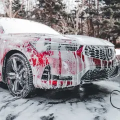 Czerwony samochód pokryty pianą, stojący na śniegu. Czy można myć auto w mrozie? Piana spływa po karoserii, tworząc ciekawe wzory.