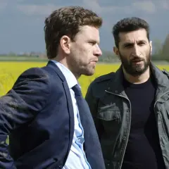 Två män står i en rapsåker, en i kostym, den andra i jacka. Scen från avdelning q filmer.