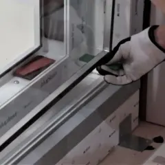 Rękawica montażysty umieszcza zieloną podkładkę pod szybę termoizolacyjną w ramie okiennej.