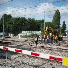 Pracownicy budowlani i dróżnik przejazdowy nadzorują prace przy torach. Koparka i ciężarówka są na miejscu.