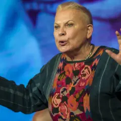 Elżbieta Dzikowska w kolorowej bluzce z haftem, gestykuluje rękami na tle niebieskiego ekranu.