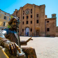 Statua in bronzo di un uomo barbuto, un monumento in Emilia Romagna, con una chiesa in mattoni sullo sfondo.