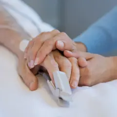 Dłoń pacjenta z pulsoksymetrem na palcu, otoczona troskliwymi dłońmi bliskiej osoby. To obraz nadziei i wsparcia na oddziale OIOM.