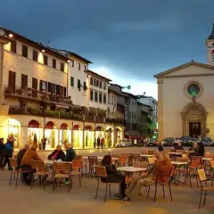 Serata in piazza a Figline Valdarno, con tavolini all'aperto e la chiesa illuminata.