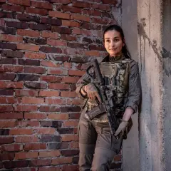 Kobieta w kamizelce Direct Action, z karabinem, opiera się o betonowy słup przy ceglanej ścianie.