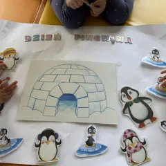 Dzieci tworzą plakat na dzień pingwina w przedszkolu, ozdabiając go naklejkami pingwinów i igloo.