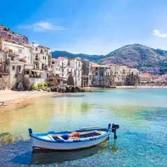 A charming fishing boat bobs in the clear turquoise water off the coast of Cefalù, a picturesque town with colorful buildings cascading down a hillside, making it one of the most beautiful places to go in Sicily.