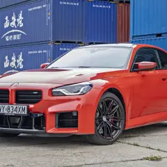 Pomarańczowe BMW M2 Competition z czarnymi felgami stoi na tle niebieskich kontenerów.