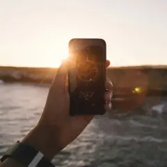 Dłoń trzyma telefon z włączonym kompasem w tle zachodzącego słońca nad morzem.