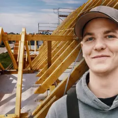 Junger Mann mit Kappe lächelt vor Dachstuhl. Ausbildung Zimmermann Gehalt ist ein guter Start.