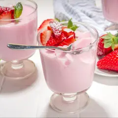 Różowa panna cotta z truskawkami, udekorowana listkami mięty i posypana cukrem pudrem. Obok świeże truskawki.