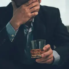 Mężczyzna w garniturze z ręką na czole, trzymający szklankę z bursztynowym płynem. Może to symbolizować problemy, stres lub nawet renta alkoholowa.