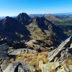 Widok z kamienistego szczytu w Tatrach Wysokich na skaliste granie, trawiaste zbocza i turkusowe jezioro.