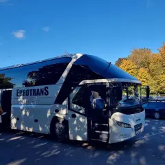 Biały autokar firmy Eurotrans z otwartymi drzwiami, zaparkowany na tle jesiennych drzew i innych samochodów.