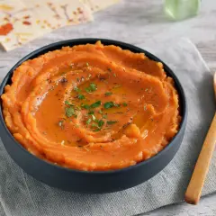 Un tazón de puré de boniato gourmet, cremoso y adornado con aceite y hierbas. Una cuchara de madera descansa a su lado.