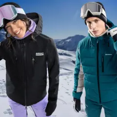 Pareja sonriente en la nieve, ella con chaqueta hardshell negra, él con chaqueta acolchada verde.