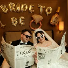 Para Młoda czyta gazetę "The Wedding Post" o miłości, przygotowując się do wesela krok po kroku. Balony "Bride to Be" i kieliszki tworzą atmosferę.