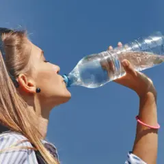 Kobieta pijąca wodę z plastikowej butelki na tle błękitnego nieba.