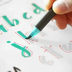 Mano practicando lettering con plantillas imprimibles gratis. Letras "a, b, c, j, i, t, u" en verde y rojo.