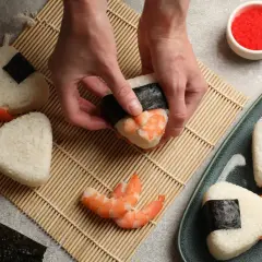 Ręce formują onigiri z krewetkami, tworząc pyszne japońskie jedzenie. Obok miska ryżu i talerz z gotowymi przysmakami.