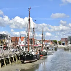 Tre segelfartyg ligger vid kaj i Husum, Tyskland. Bakom dem syns en rad hus och en bro över kanalen.