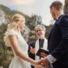 Młoda para trzymająca się za ręce podczas ceremonii ślubnej w plenerze. Celebrans z uśmiechem udziela im piękne błogosławieństwo.
