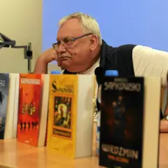 Mężczyzna zamyślony nad stosem książek, wśród nich "Wiedźmin" i inne tytuły Sapkowskiego, idealne dla fanów szukających książek podobnych do Wiedźmina.