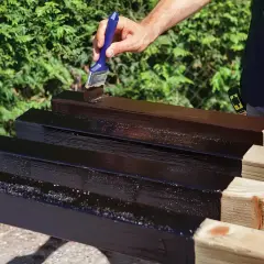 Mano che applica vernice scura su listelli di legno grezzo, un passo essenziale per imparare come impermeabilizzare il legno e proteggerlo.