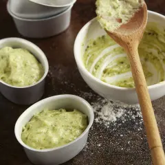 Kleine Förmchen werden mit einer grünen Masse gefüllt, die nach Pistaziencreme selber machen aussieht. Ein Holzlöffel liegt in einer Schüssel mit der Masse.