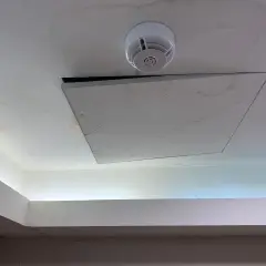 Un soffitto in cartongesso con una botola aperta, un rilevatore di fumo e macchie d'acqua. Il montaggio botola cartongesso è visibile.