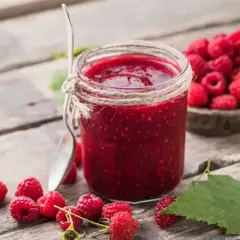 Słoik domowego sosu malinowego, otoczony świeżymi malinami i liśćmi, na drewnianym stole. Idealny dodatek do deserów.