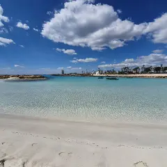 Kristallklart vatten och vit sand vid Nissi Beach, Cypern. Små båtar guppar i det turkosa vattnet under en blå himmel med vita moln.
