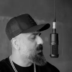 Artysta w czapce z daszkiem i łańcuchem na szyi nagrywa utwór rnb przy mikrofonie.