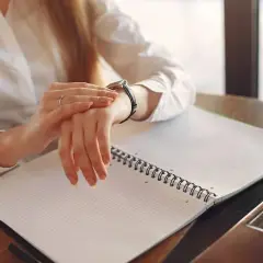 Kobieta w białej koszuli sprawdza zegarek, pracując po godzinach. Na biurku leży otwarty notatnik.