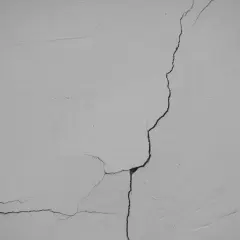 Widoczne pękanie ścian w narożnikach, tworzące sieć drobnych i większych szczelin na jasnej, tynkowanej powierzchni.