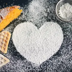A slice of cake, heart-shaped cookies, and a heart shape made of powdered sugar on a dark surface.