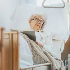 Ältere Dame liest im Bett, eine Auszeit in der Kurzzeitpflege ohne Pflegegrad. Gemütlich eingekuschelt mit einem Buch.