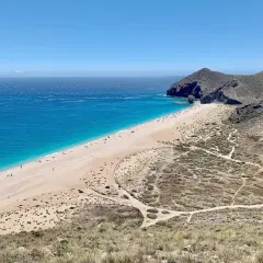 Una cala de arena dorada se extiende junto a un mar turquesa. Senderos serpentean por las colinas áridas que rodean la playa.