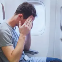 Mężczyzna w samolocie z rękami na twarzy, wyraźnie cierpiący na jet lag po długiej podróży.