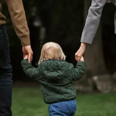 Dziecko z blond włosami, w zielonej kurtce, trzyma za ręce rodziców. To obraz przysposobienia niepełne, pełnego miłości i nadziei.