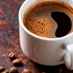 En vit kopp med mörkt kaffe och skum. Kaffebönor ligger utspridda. Undrar du hur mycket koffein i en kopp kaffe?