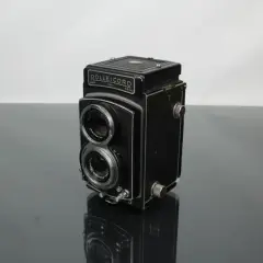 A vintage Rolleicord camera sits on a reflective surface, its twin lenses and classic design hinting at a rich photographic history.