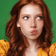 A young woman with fiery red hair, often symbolizing passion and uniqueness, puckers her lips against a vibrant green backdrop.