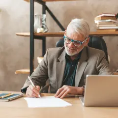 Homme âgé souriant écrivant sur un papier à son bureau, avec un ordinateur portable et une plante.