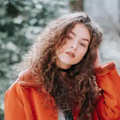 Joven con melenas rizadas y un abrigo naranja, posando con confianza.